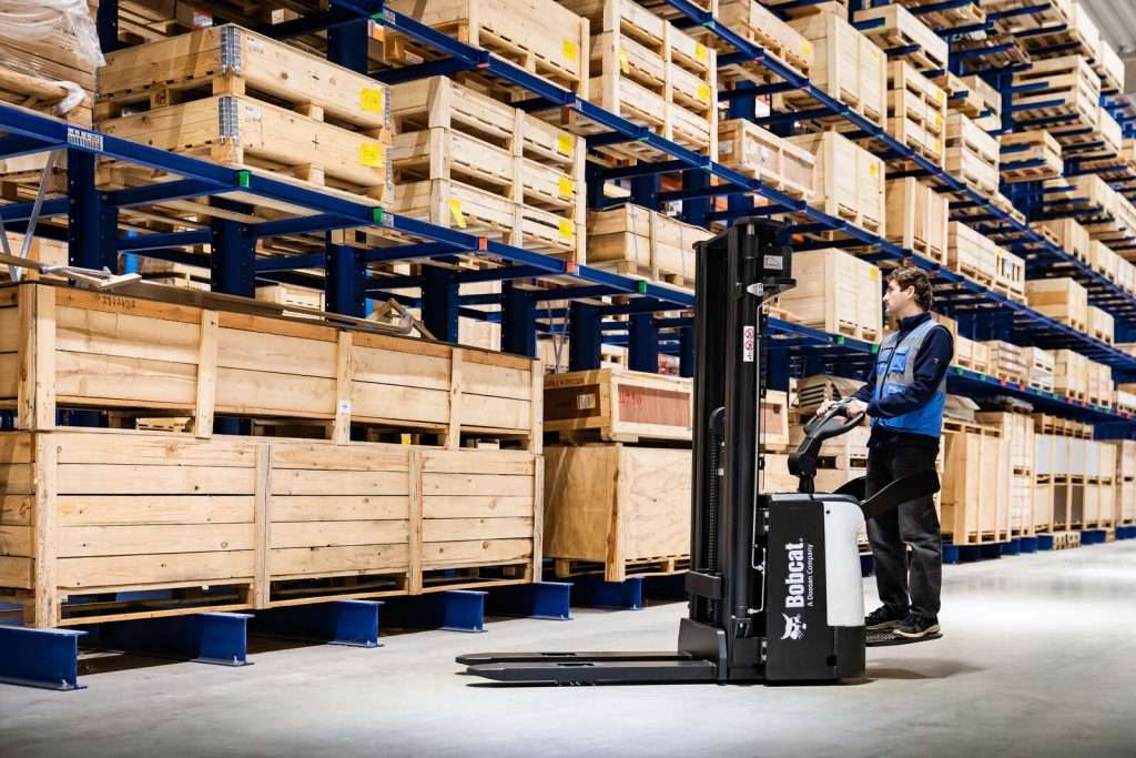 Mitarbeiter bedient Hochhubwagen zwischen hohen Holzregalen